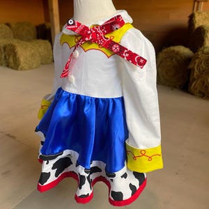May include: A white, blue, and yellow cowgirl costume with a red bandana bow. The dress features a white top with yellow accents, a blue skirt, and a cow-print hem with red trim. The sleeves have yellow cuffs.
