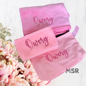 May include: A set of three pink items with the name "Qwery" embroidered on each. The set includes a headband, a small pouch, and a larger towel. The items are all made of a soft, plush fabric.