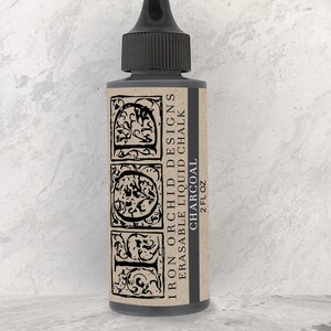 May include: A charcoal-colored liquid chalk bottle with a black cap. The label reads "Iron Orchid Designs Erasable Liquid Chalk Charcoal 2 fl oz" with decorative scrollwork. The bottle is on a light gray background.