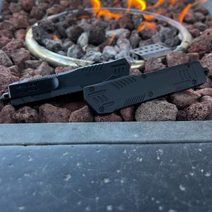 May include: Two black knives with textured handles are displayed on a bed of reddish-brown lava rocks. The knives are positioned near a fire pit with flames visible in the background. The knives have a sleek, modern design.