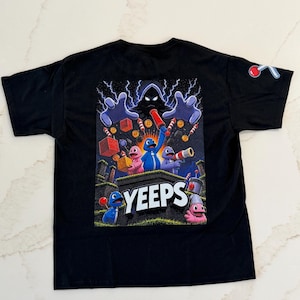 May include: A black short-sleeve t-shirt featuring a large graphic print on the back. The design displays colorful cartoon characters, the text YEEPS, and a dark hooded figure with lightning. A small icon of a red and white tool is on the sleeve.