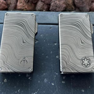 May include: Two rectangular, silver-colored metal cases with a wave-like pattern. One case features a Mandalorian symbol, and the other has an Imperial symbol. Both have a small, hinged top and rounded corners.