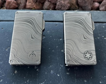 May include: Two rectangular, silver-colored metal cases with a wave-like pattern. One case features a Mandalorian symbol, and the other has an Imperial symbol. Both have a small, hinged top and rounded corners.