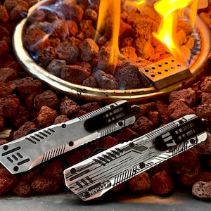 May include: Two silver pocket knives with black clips and intricate designs are displayed on a bed of reddish-brown rocks. One knife has the text "HN-117". The knives are near a fire pit with flames.