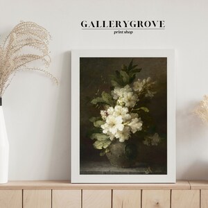 May include: A framed print of a still life painting featuring a bouquet of white flowers in a vase. The flowers are arranged in a cluster with green leaves and are set against a dark background. The print is in a white frame and is displayed on a wooden shelf.