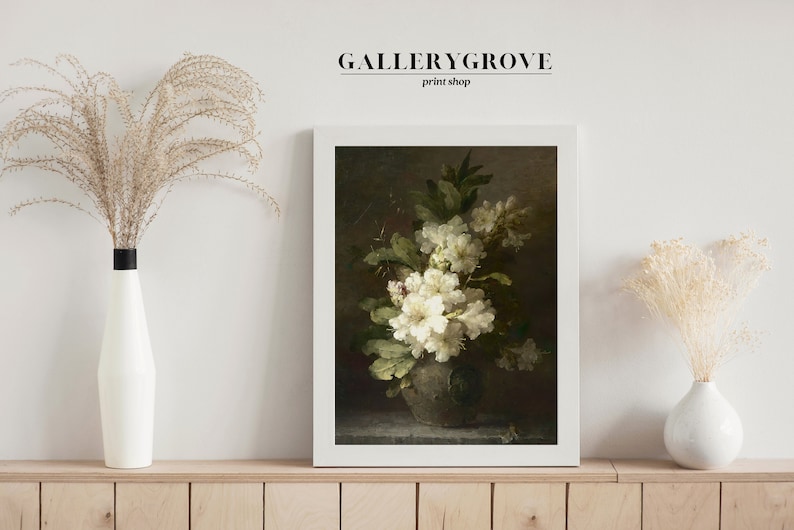 May include: A framed print of a still life painting featuring a bouquet of white flowers in a vase. The flowers are arranged in a cluster with green leaves and are set against a dark background. The print is in a white frame and is displayed on a wooden shelf.