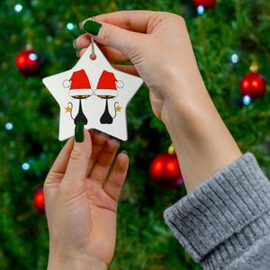 Op de afbeelding: Een witte, stervormige kerstversiering met een grillig ontwerp van twee zwarte kat silhouetten met rode kerstmutsen. De versiering heeft een gouden touwtje om op te hangen, tegen een vage kerstboom achtergrond.