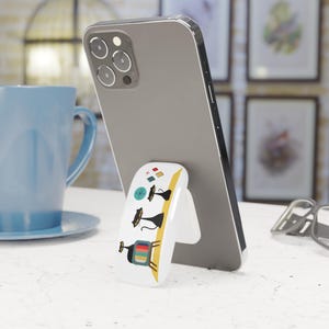 May include: A white phone stand with a colorful design featuring a retro television and three black cats. The stand is designed to prop up a smartphone.