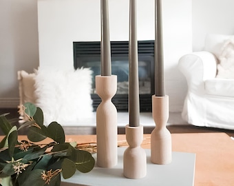 May include: Three grey taper candles in wooden candle holders on a grey book with the word "HOME" printed on the cover. The candles are on a wooden table with a green plant in the foreground.