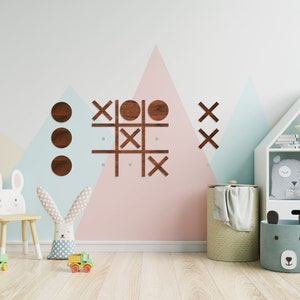 Pu&ograve; includere: Un gioco di tris in legno con cerchi e croci in finitura noce. Il gioco &egrave; montato su una parete in una cameretta con uno schema di colori blu chiaro, rosa e beige. La stanza presenta anche una libreria bianca con una stella e un peluche a forma di nuvola, un cavallo a dondolo bianco e uno sgabello bianco con un coniglio sopra.