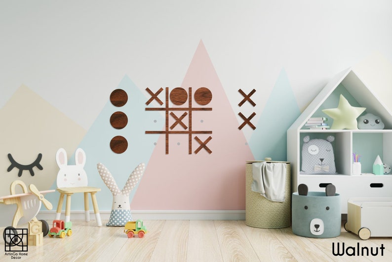 Pu&ograve; includere: Un gioco di tris in legno con cerchi e croci in finitura noce. Il gioco &egrave; montato su una parete in una cameretta con uno schema di colori blu chiaro, rosa e beige. La stanza presenta anche una libreria bianca con una stella e un peluche a forma di nuvola, un cavallo a dondolo bianco e uno sgabello bianco con un coniglio sopra.