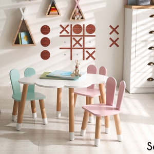 Pu&ograve; includere: Un tavolo bianco a forma di nuvola con quattro sedie in legno. Due sedie sono di colore azzurro con orecchie di coniglio e due sedie sono di colore rosa con orecchie di coniglio. Le sedie hanno i piedi dipinti di bianco. Il tavolo ha quattro gambe in legno con i piedi dipinti di bianco. Il tavolo &egrave; posizionato in una sala giochi con una cassettiera bianca e una parete decorata con forme geometriche in legno.