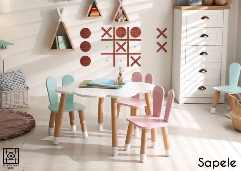 Pu&ograve; includere: Un tavolo bianco a forma di nuvola con quattro sedie in legno. Due sedie sono di colore azzurro con orecchie di coniglio e due sedie sono di colore rosa con orecchie di coniglio. Le sedie hanno i piedi dipinti di bianco. Il tavolo ha quattro gambe in legno con i piedi dipinti di bianco. Il tavolo &egrave; posizionato in una sala giochi con una cassettiera bianca e una parete decorata con forme geometriche in legno.