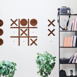 Pu&ograve; includere: Un gioco di tris in legno su una parete bianca. I pezzi del gioco sono cerchi e croci in finitura noce. Il gioco &egrave; parzialmente giocato con tre cerchi e tre croci. La parete &egrave; decorata con due piante in vaso.