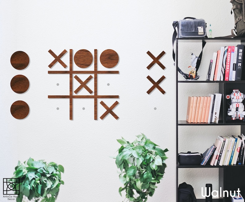 Pu&ograve; includere: Un gioco di tris in legno su una parete bianca. I pezzi del gioco sono cerchi e croci in finitura noce. Il gioco &egrave; parzialmente giocato con tre cerchi e tre croci. La parete &egrave; decorata con due piante in vaso.