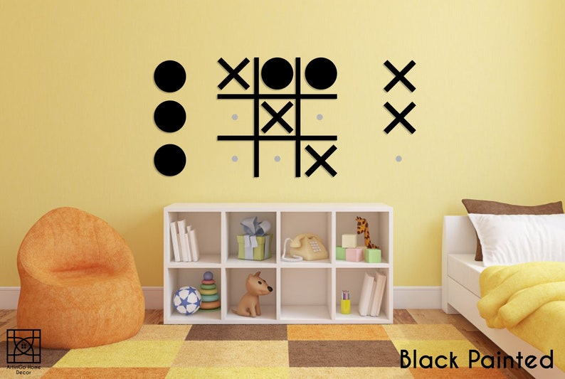 Pu&ograve; includere: Gioco di tris in legno dipinto di nero con cerchi e croci su una parete gialla in una cameretta. Il gioco &egrave; montato sulla parete sopra una libreria bianca con giocattoli e libri.