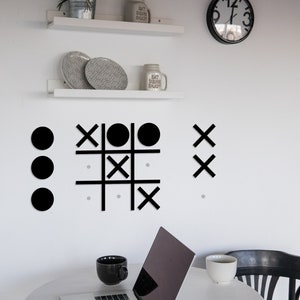 Pu&ograve; includere: Gioco di tris in legno dipinto di nero con cerchi neri e X su una parete bianca. Nell'immagine sono visibili anche un computer portatile, due tazze e un orologio.