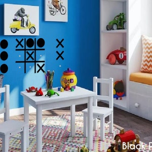 Pu&ograve; includere: Un set di tavolo e sedie bianche per bambini, con un gioco di tris nero sul muro blu dietro. Ci sono giocattoli sul tavolo e un cestino sul pavimento.