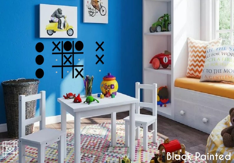 Pu&ograve; includere: Un set di tavolo e sedie bianche per bambini, con un gioco di tris nero sul muro blu dietro. Ci sono giocattoli sul tavolo e un cestino sul pavimento.