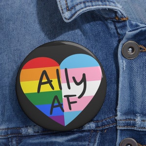 May include: A black button pin with a heart design. The left side of the heart is a rainbow, and the right side features the transgender flag. The words "Ally AF" are written in black script across the heart.