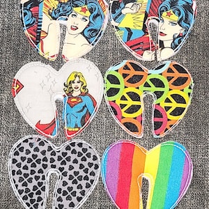 May include: Six heart-shaped fabric patches with different designs. The designs include Wonder Woman, Superman, peace signs, hearts, and a rainbow pattern.
