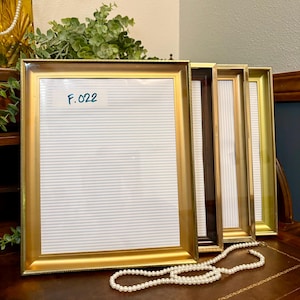 May include: Four rectangular picture frames with gold and brown finishes are displayed. The frames have a variety of styles, including a gold frame with the text "F.022" on the glass. A pearl necklace is draped in front of the frames.