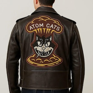 Può includere: Giacca in pelle marrone con una grande toppa sul retro raffigurante un gatto dei cartoni animati con la scritta "ATOM CATS". La toppa ha una combinazione di colori marrone, giallo e nero. La giacca ha un classico stile biker.