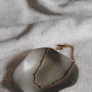 May include: A delicate gold necklace with a beaded chain rests on a small, gray, organically shaped dish. The dish sits on a textured, off-white linen surface. The necklace has a simple, elegant design.