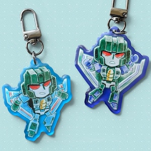May include: Two keychains with a cartoon robot design. The robots are coloured green and blue with red eyes, attached to silver key rings. The keychains are set against a light blue background.