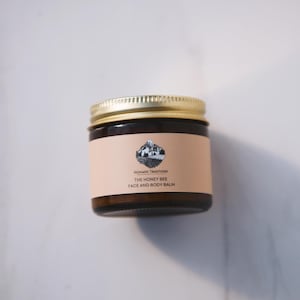 May include: A brown glass jar with a gold lid containing a face and body balm. The label is peach colored with black text that reads "NOMADIC TRADITIONS THE HONEY BEE FACE AND BODY BALM".