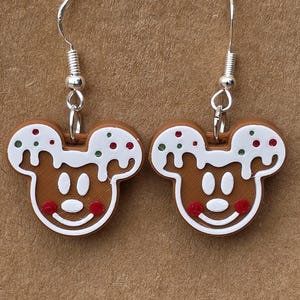 May include: A pair of novelty earrings shaped like gingerbread Mickey Mouse faces. The earrings are brown with white icing and red and green sprinkles. The earrings have silver-coloured fish hook ear wires.