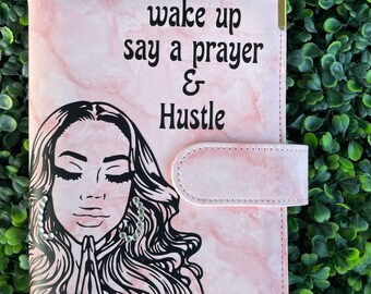 Peut inclure: Un agenda en simili cuir rose avec un motif marbré et une illustration noire d'une femme avec les mains jointes en prière. Le texte "wake up say a prayer & Hustle" est imprimé sur le devant.