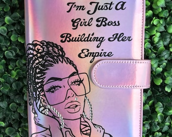 Peut inclure: Un classeur rose iridescent avec une illustration en noir et blanc d'une femme portant des lunettes et des boucles d'oreilles. Le texte "I'm Just A Girl Boss Building Her Empire" est imprimé sur le devant.
