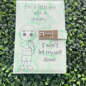 Puede incluir: Un diario de color verde y blanco con un patrón de mármol, una cerradura y una ilustración de un niño de dibujos animados. El diario tiene el texto "I'm a little guy with a dream... I won't let myself down."