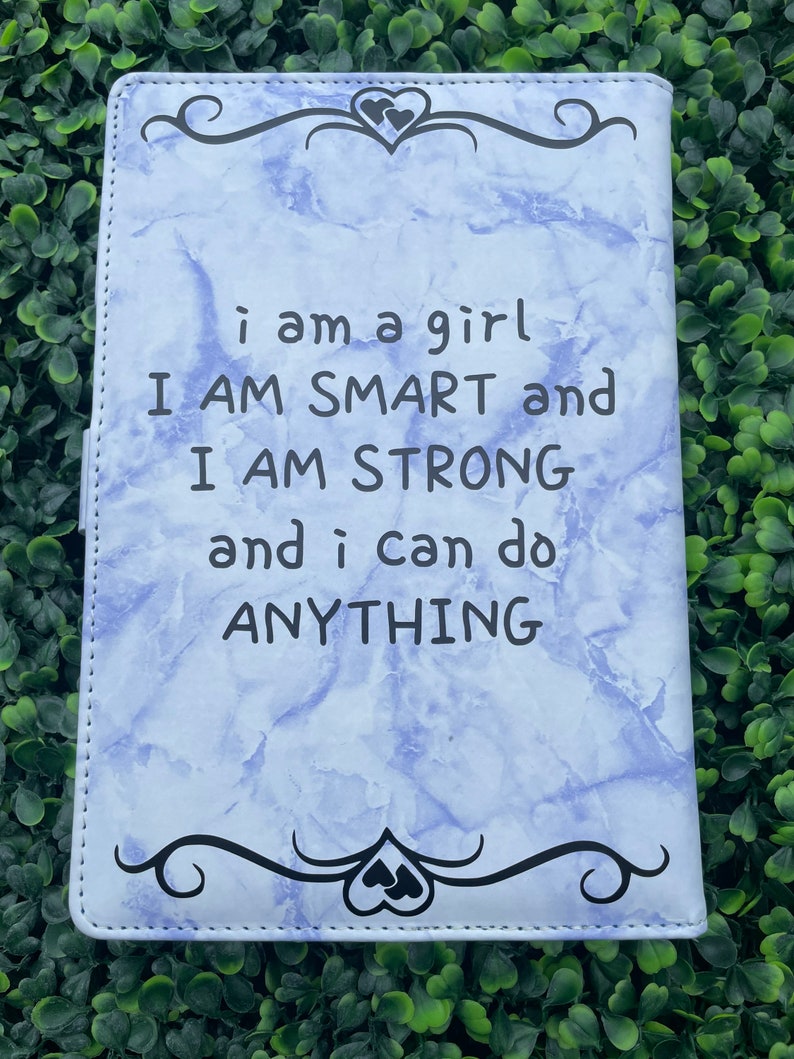 May include: A light blue notebook with a marble pattern and black text that reads "i am a girl I AM SMART and I AM STRONG and i can do ANYTHING". The notebook has a black decorative border with two hearts at the top and bottom.