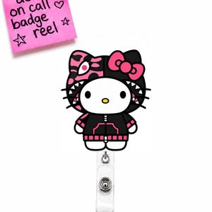 Puede incluir: Un portacredenciales con un personaje de dibujos animados de diseño dividido, un lado negro y otro rosa con estampado de leopardo. El personaje lleva una sudadera negra con detalles rosas y un lazo rosa. Una nota rosa dice doll on call badge reel.