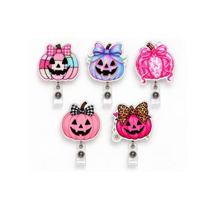 May include: Five badge reels featuring jack-o'-lantern pumpkin designs in various shades of pink. Each pumpkin has a unique pattern, including plaid, leopard print, and ribbon motifs, topped with decorative bows and attached to metal clips.