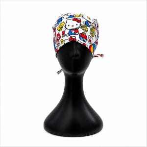 Op de afbeelding: Een witte operatiemuts met een kleurrijk herhalend patroon van Hello Kitty-figuren, rode appels, blauwe broodtrommels, gele eekhoorns en rode tulpen. De muts is voorzien van een striksluiting aan de achterzijde voor een goede pasvorm.
