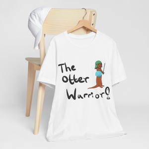 May include: White t-shirt with the text "The Otter Warrior!" in black script. The shirt features a cartoon otter wearing a green helmet and holding a sword and a blue shell. A white baseball cap rests on a wooden chair.