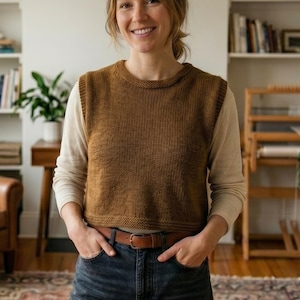 May include: A woman wearing a brown knitted vest over a cream-colored long-sleeved shirt and blue jeans. The room has bookshelves, a leather armchair, and a patterned rug. A large potted plant is in the foreground.
