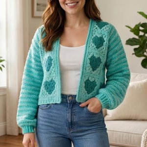 May include: A turquoise crochet cardigan with heart-shaped designs. The open-front sweater has long sleeves and is paired with blue jeans. The cardigan is a light, airy garment, perfect for layering.