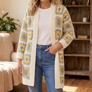 Puede incluir: Un cárdigan largo color crema con un patrón geométrico en amarillo y azul. El suéter abierto se usa sobre una camiseta blanca y unos vaqueros azules. El cárdigan llega a la rodilla y tiene un corte holgado.