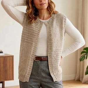 May include: A beige knit vest with a ribbed design, worn over a white long-sleeved shirt. The vest has a round neckline and a vertical opening. The person is wearing gray pants and a brown belt.