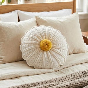 Può includere: Un letto è fatto con lenzuola e cuscini color crema. Un cuscino decorativo rotondo all'uncinetto bianco con un centro giallo è appoggiato sopra. Una coperta in maglia grigia è ai piedi del letto. La testiera è in legno chiaro.