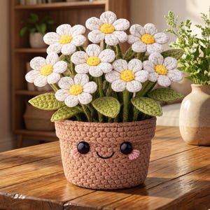 May include: A crocheted daisy flower arrangement in a brown pot with a smiling face. The daisies have white petals and yellow centres. The pot is on a wooden table. A vase with greenery is next to the flower arrangement.
