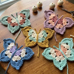 May include: A collection of colorful, crocheted butterfly appliques in various shades of blue, green, yellow, and purple, with cream and peach accents. The butterflies are arranged on a round wooden table, along with wooden beads and a pair of scissors.