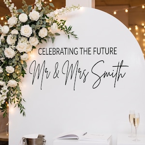 May include: A white, arched wedding sign with the text "CELEBRATING THE FUTURE" and "Mr & Mrs Smith" in elegant script. The sign is adorned with white roses and greenery. Champagne glasses and a guest book are visible.