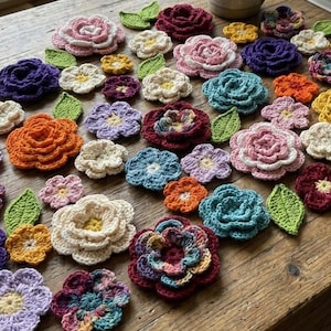 May include: A collection of colorful, crocheted flowers and leaves arranged on a wooden table. The flowers are in various sizes and shades, including pink, purple, orange, yellow, and blue. Green leaves are interspersed among the flowers.