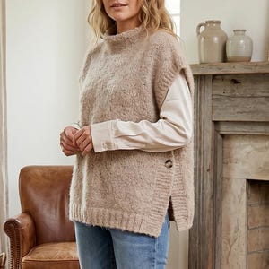 Puede incluir: Una mujer con un chaleco de punto beige sobre una camisa blanca de manga larga y vaqueros azules. El chaleco tiene cuello alto y botones laterales. Está en una habitación con chimenea y alfombra.