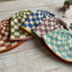 May include: Five handmade crochet beanies in various colors, including orange, green, blue, purple, and turquoise. Each beanie features a checkered pattern and a contrasting brim. The beanies are displayed on a wooden surface.